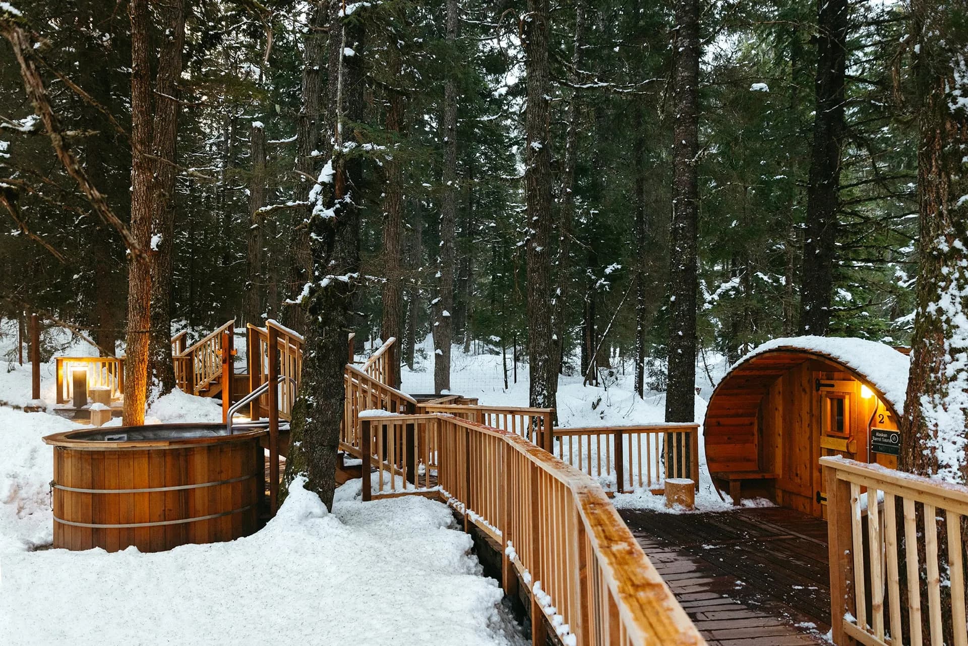 Snow-covered forest loop with barrel sauna and wooden soaking tub along a boardwalk at Alyeska Nordic Spa
