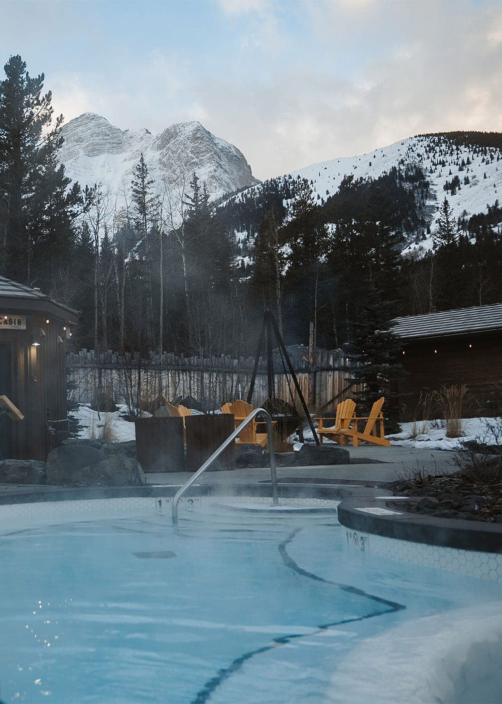 Steaming outdoor hot pool with snow-capped Rocky Mountain peaks and Adirondack chairs at Kananaskis Nordic Spa
