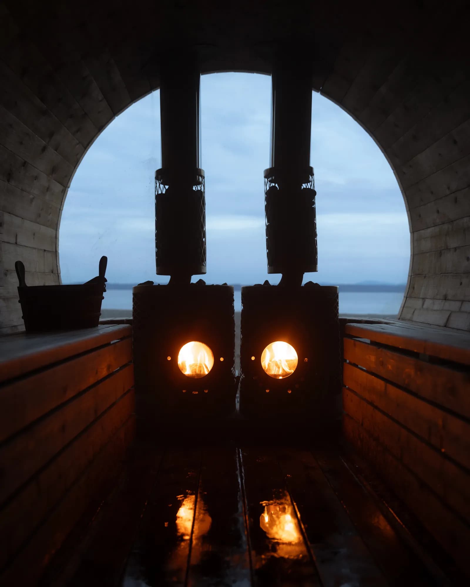 Bywater Sauna barrel interior with wood-burning stoves and round window at dusk