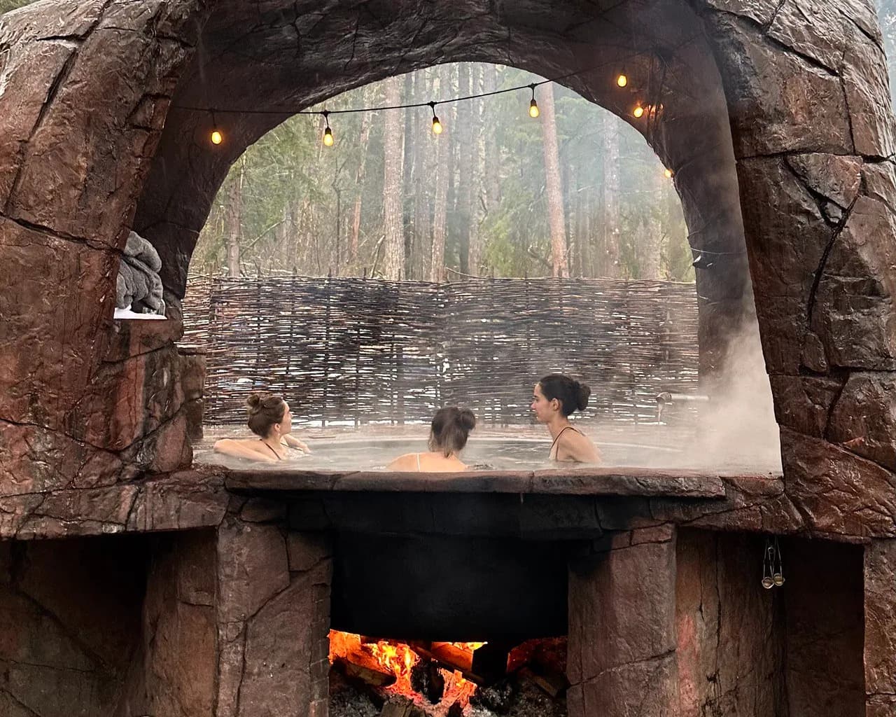 Guests soaking in the Cannibal Hot Tub, a large iron kettle heated over an open wood fire under a stone arch in the forest