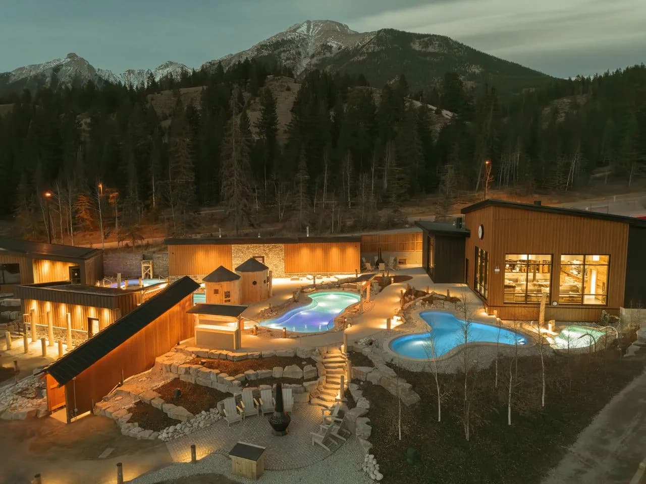 Aerial view of Everwild Canmore Nordic Spa showing outdoor hot pools, barrel saunas, and lodge buildings with Rocky Mountain backdrop at dusk