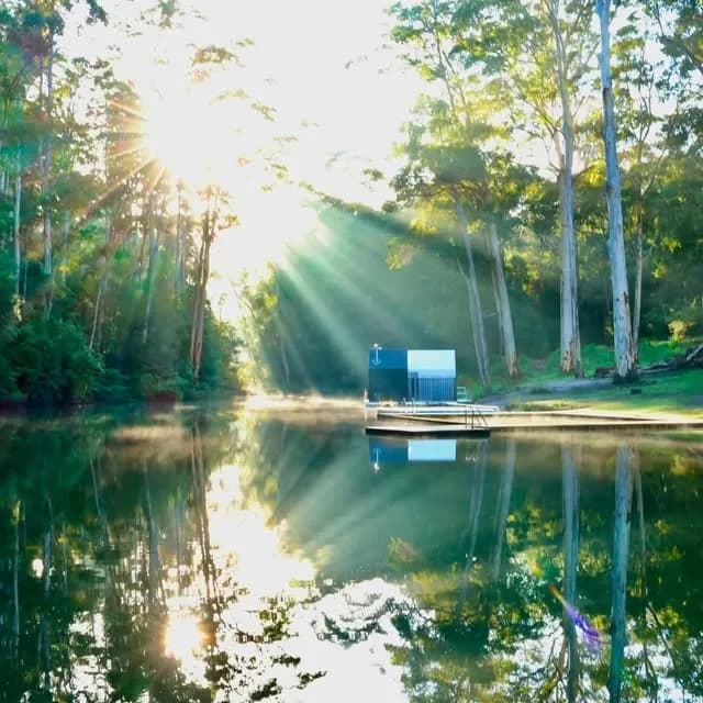 The Floating Sauna moored on Pemberton Pool at sunrise, surrounded by tall karri forest with sun rays through the trees