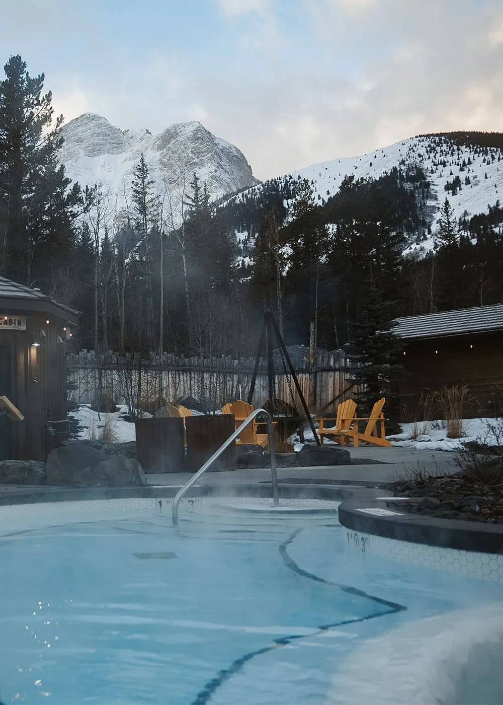 Steaming outdoor hot pool with snow-capped Rocky Mountain peaks and Adirondack chairs at Kananaskis Nordic Spa