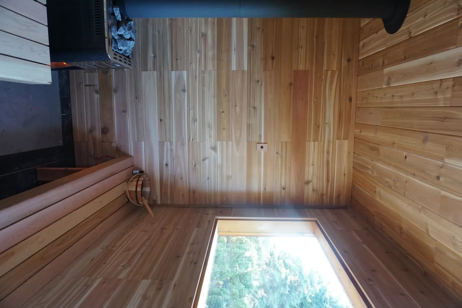 Cedar-lined interior of the Prana Sauna with bench seating, wood-fired stove, and forest view through the window