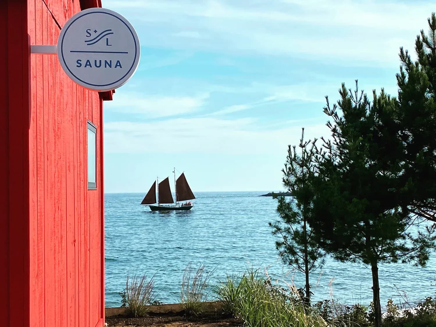 The red Sisu + Löyly sauna building with its S+L sign on the shore of Lake Superior in Grand Marais, a sailboat passing on the water