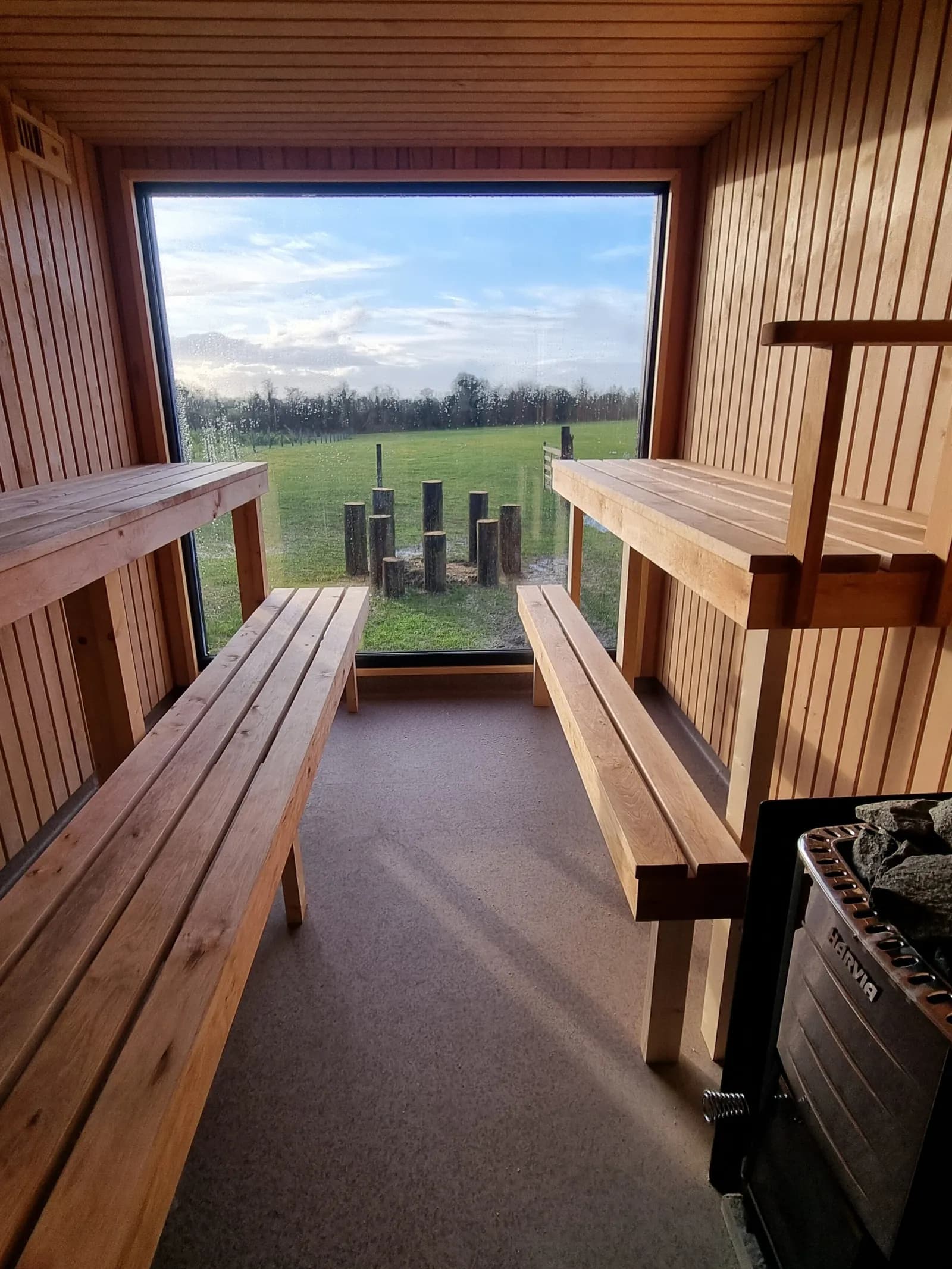 Interior of The Goat House Sauna showing wood-lined benches, Harvia wood burner, and large window overlooking green Irish countryside