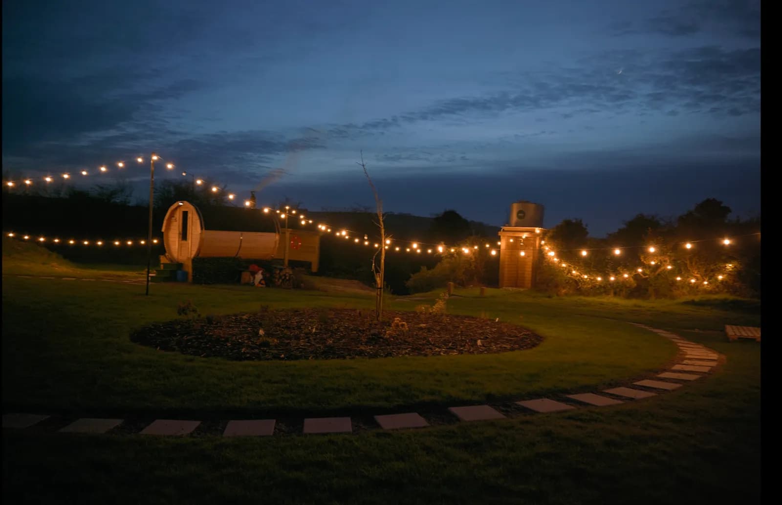 Evening view of Hidden Sauna Bridgefield with string lights illuminating the barrel sauna and fire tower against a twilight sky