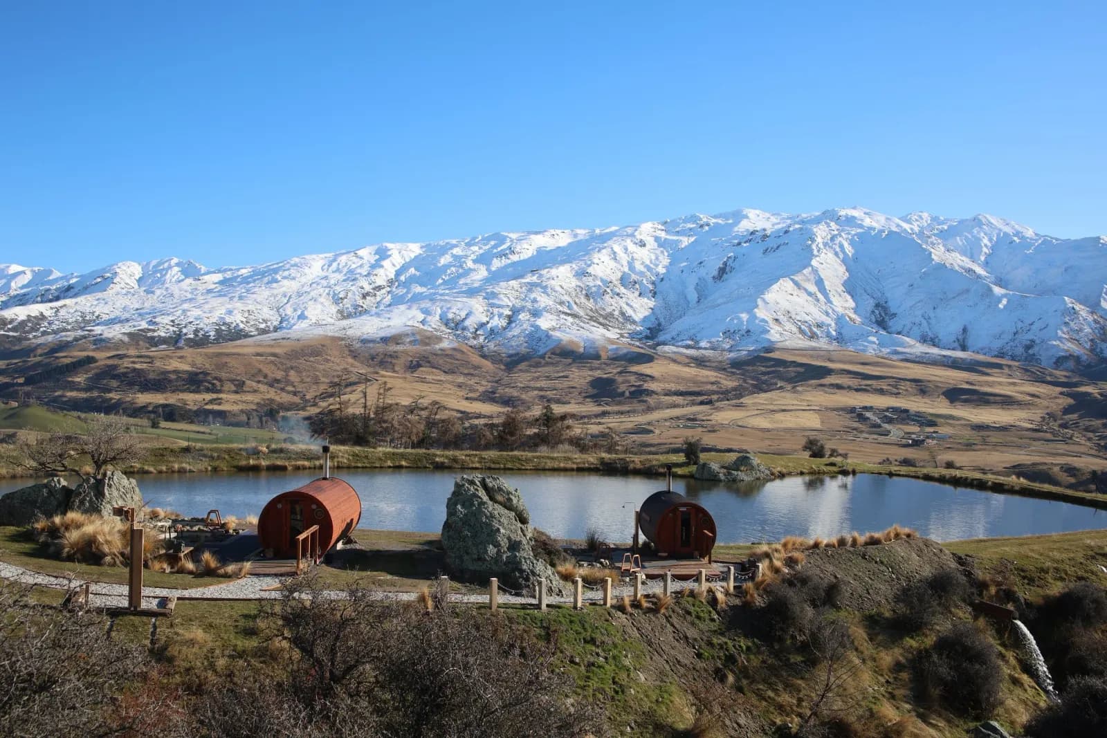 Two cedar barrel saunas beside a cold-plunge pond with snow-capped Cardrona mountains behind, at The Secret Sauna Cardrona