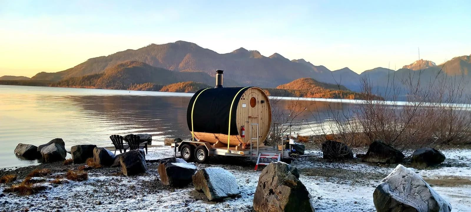 Cedar barrel sauna on a trailer beside the ocean at sunset with snow-dusted mountains near Ucluelet
